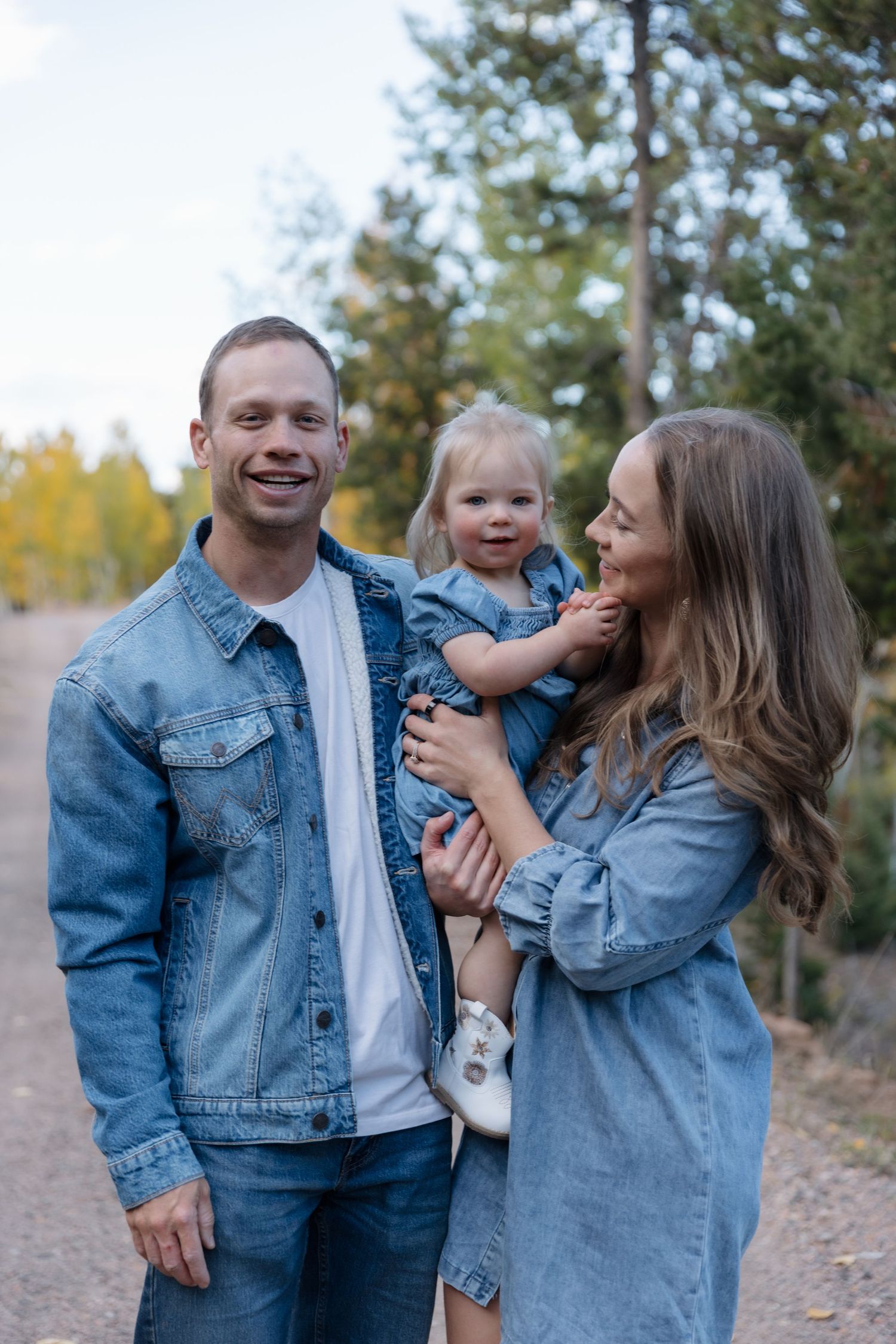 Smiling family in matching denim outfits holding toddler on a tree-lined path in autumn.
