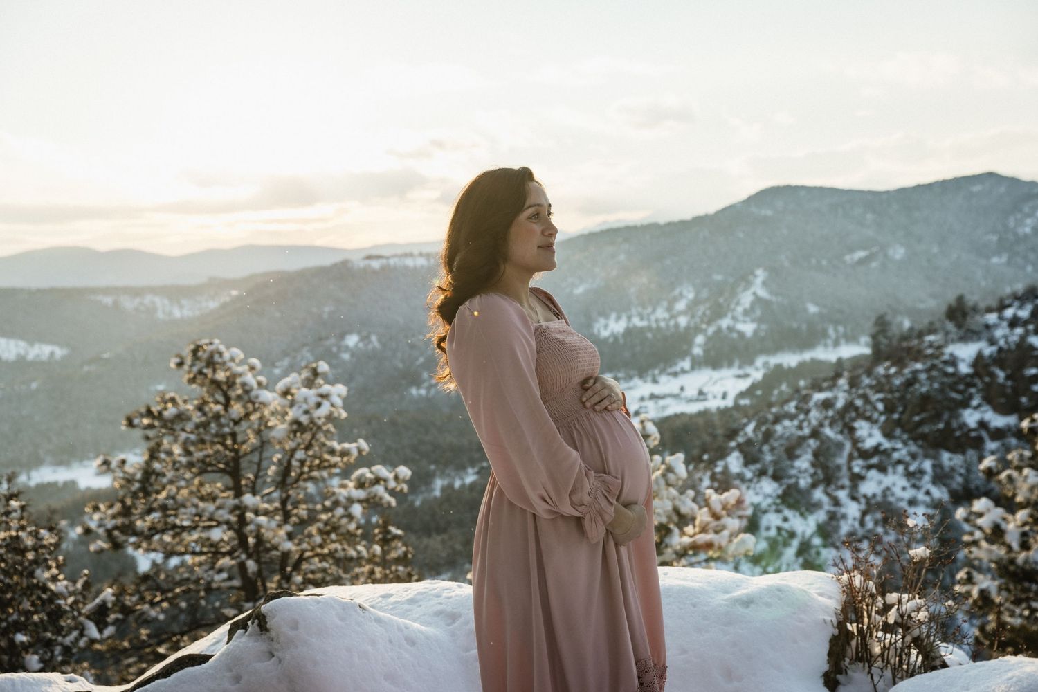 Pregnant woman in pink dress cradling baby bump at golden sunset over snowy mountains.