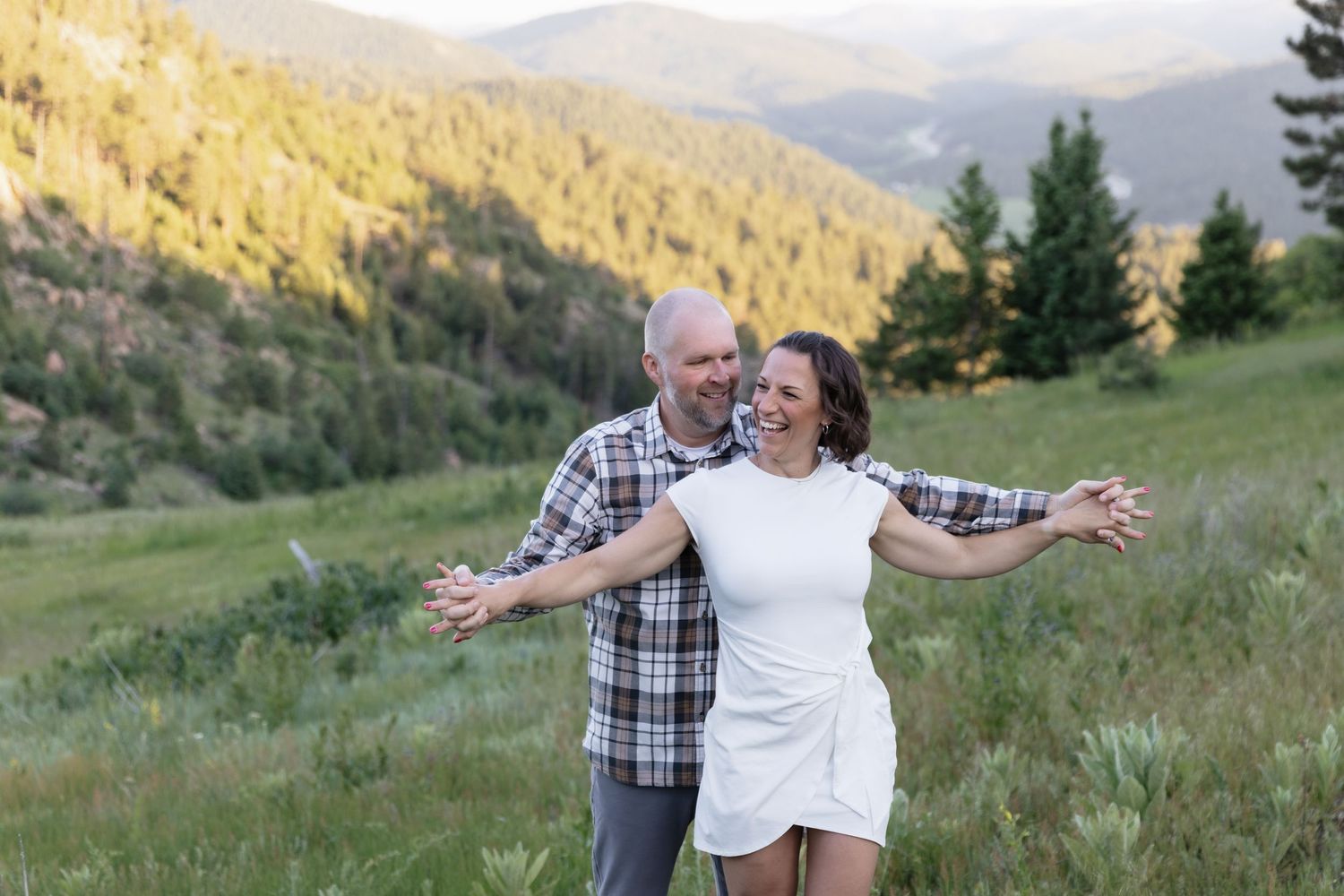 Happy couple posing in a lush green mountain valley with pine forest backdrop.