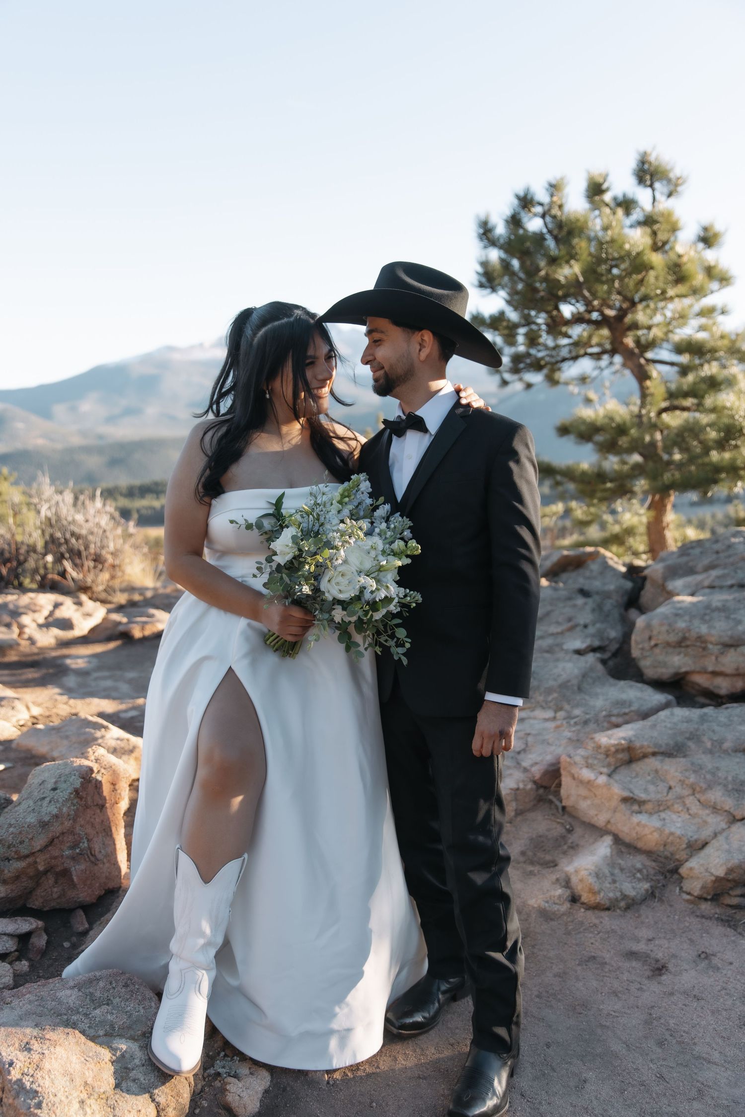 Couple in western-style wedding attire embracing on rocky mountain overlook at golden hour.