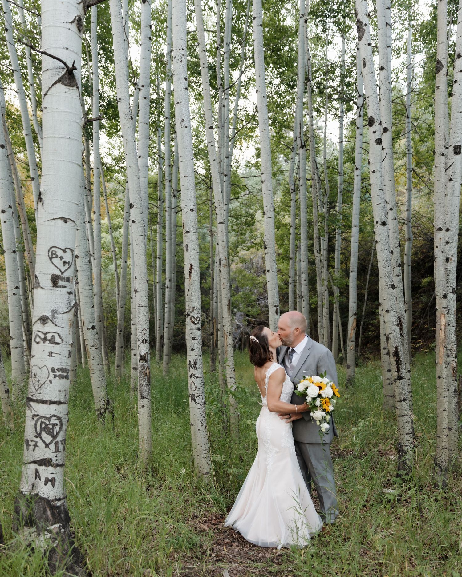 Bride and groom sharing a kiss among white aspen trees with carved hearts in a forest setting.