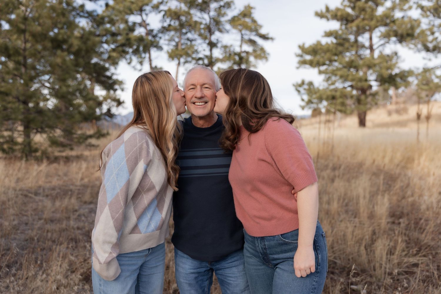Two daughters kissing their laughing father on the cheeks outdoors in a field.