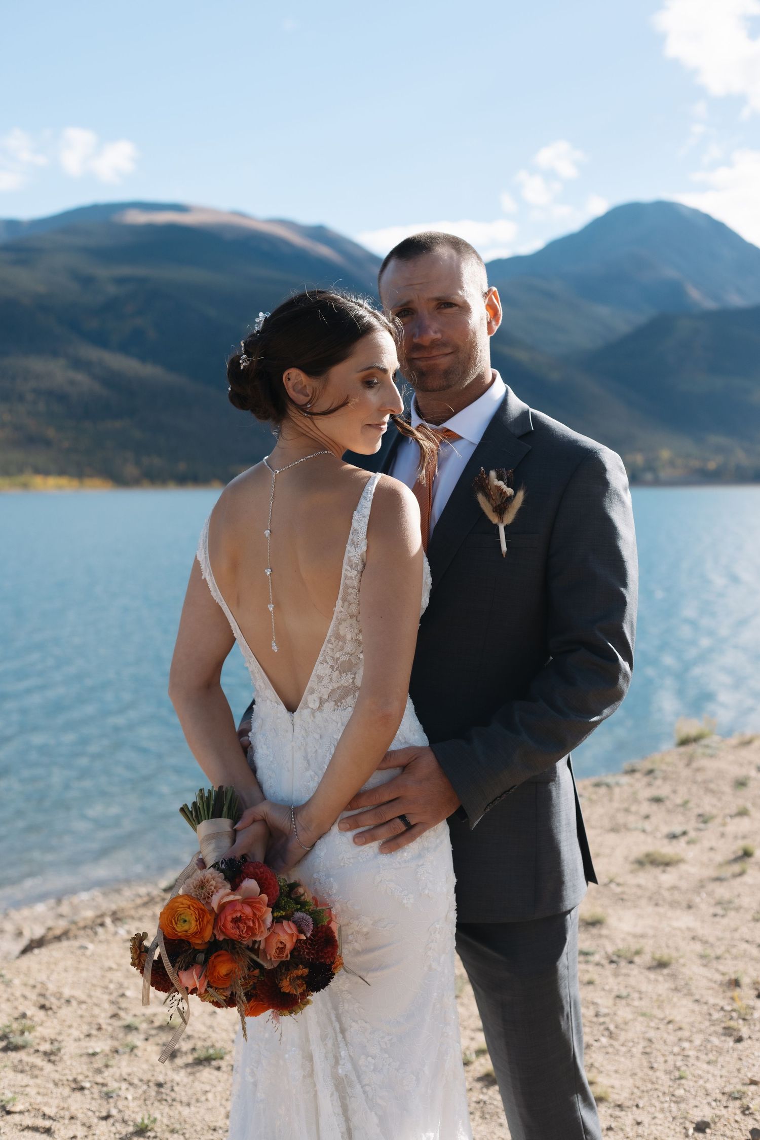 Bride and groom portrait by a blue mountain lake, bride holding vibrant orange bouquet in lace gown. Twin Lakes