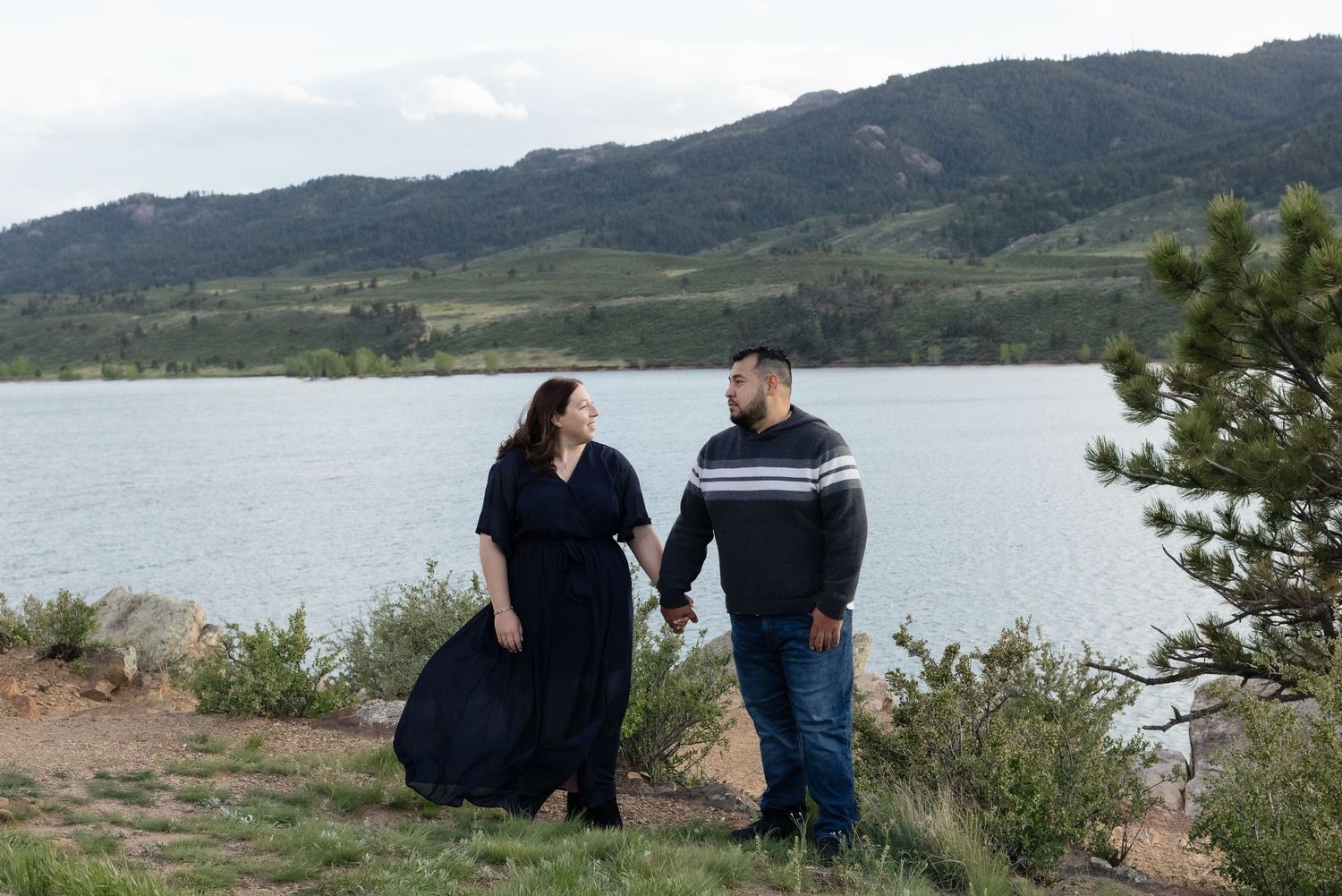 Couple holding hands by a mountain lake, woman in navy dress, man in striped sweater and jeans, green hills backdrop.