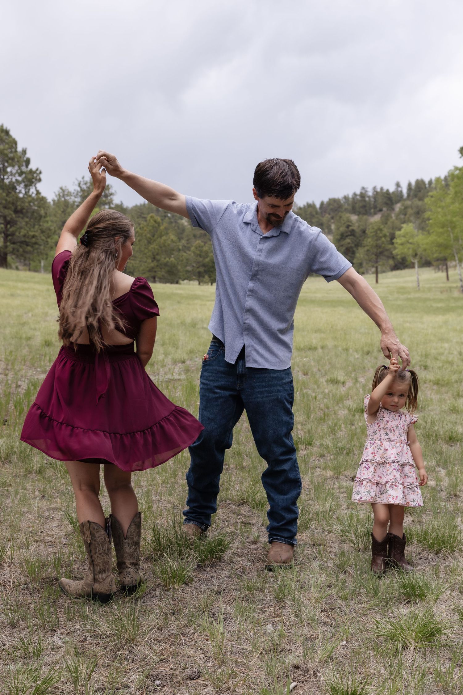 A family of three dancing in a meadow, parents spinning their young daughter in cowboy boots and summer dresses.