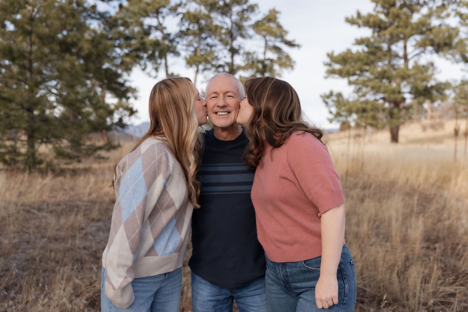 Two daughters kissing their laughing father on the cheeks in a sunny pine forest.