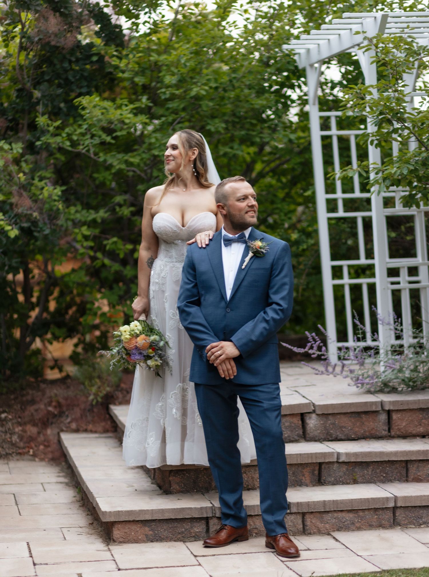 Tall bride in white gown and groom in blue suit pose in garden.