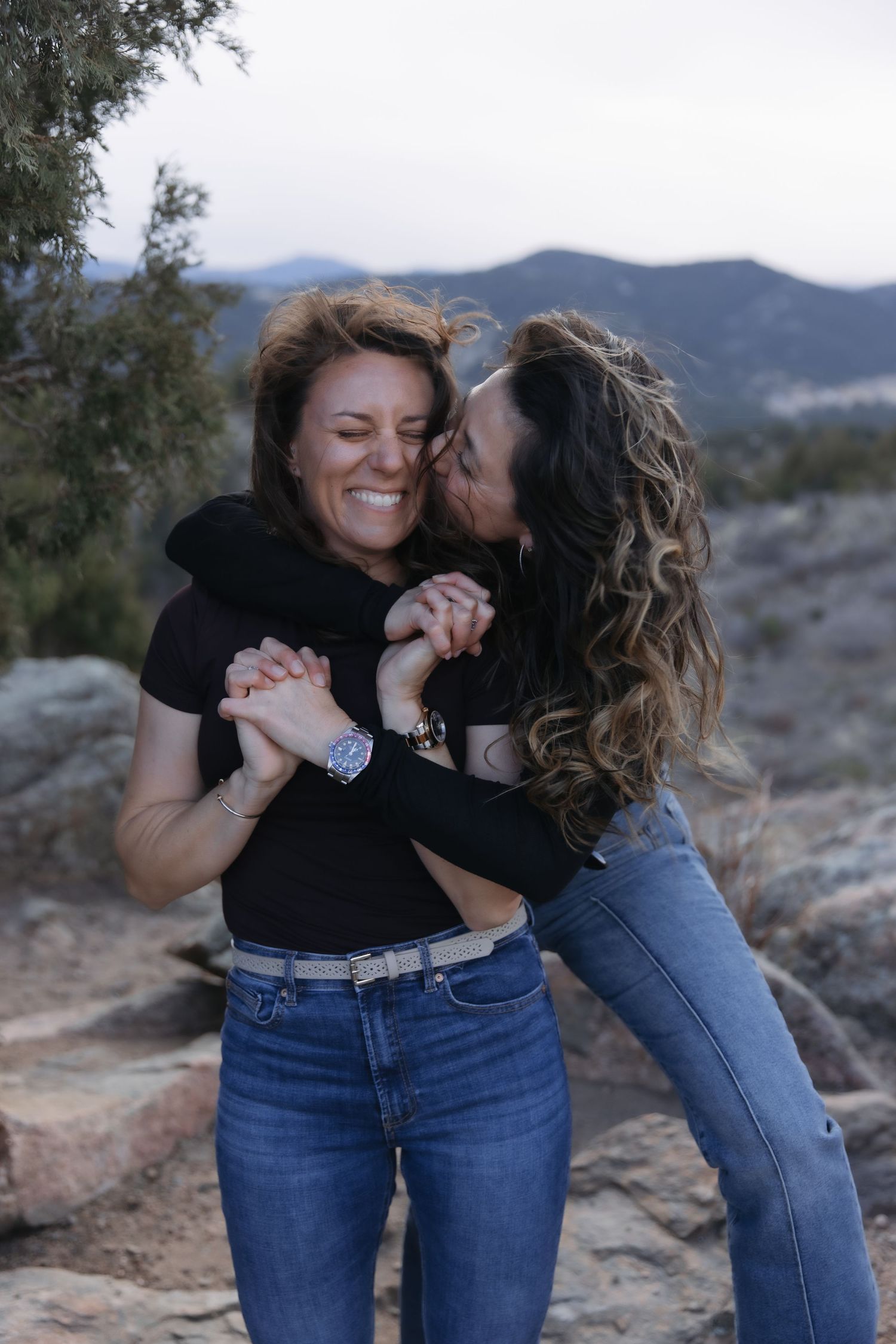 Two women embracing and laughing together on rocky terrain with mountain landscape in background.