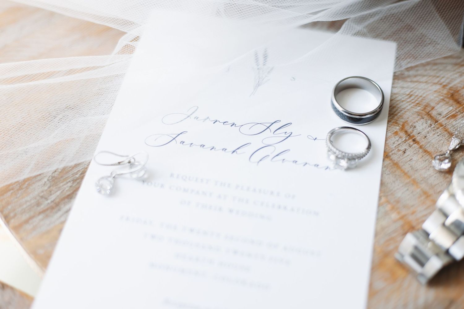 Wedding invitation with elegant calligraphy script displayed alongside diamond rings and silver wedding bands on wood surface.