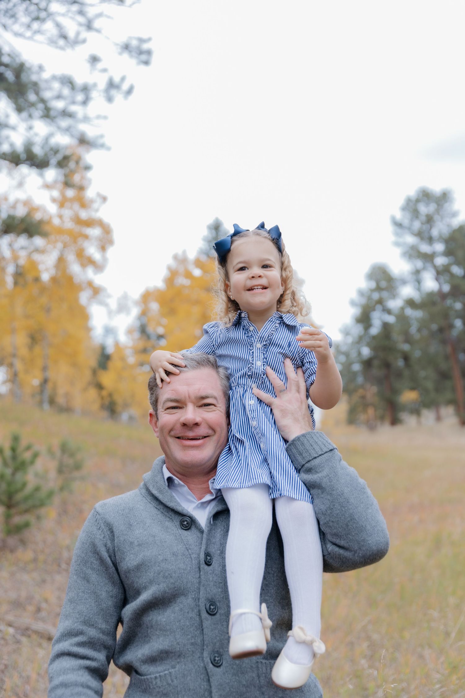 Smiling man carrying a young girl in a blue dress on his shoulders in an autumn forest setting.
