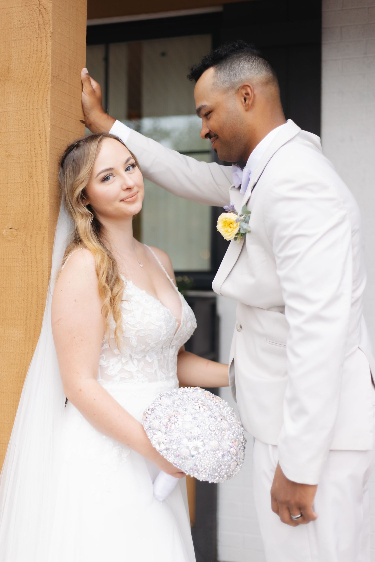 Bride in lace gown holding crystal bouquet with groom in white suit beside a wooden building.