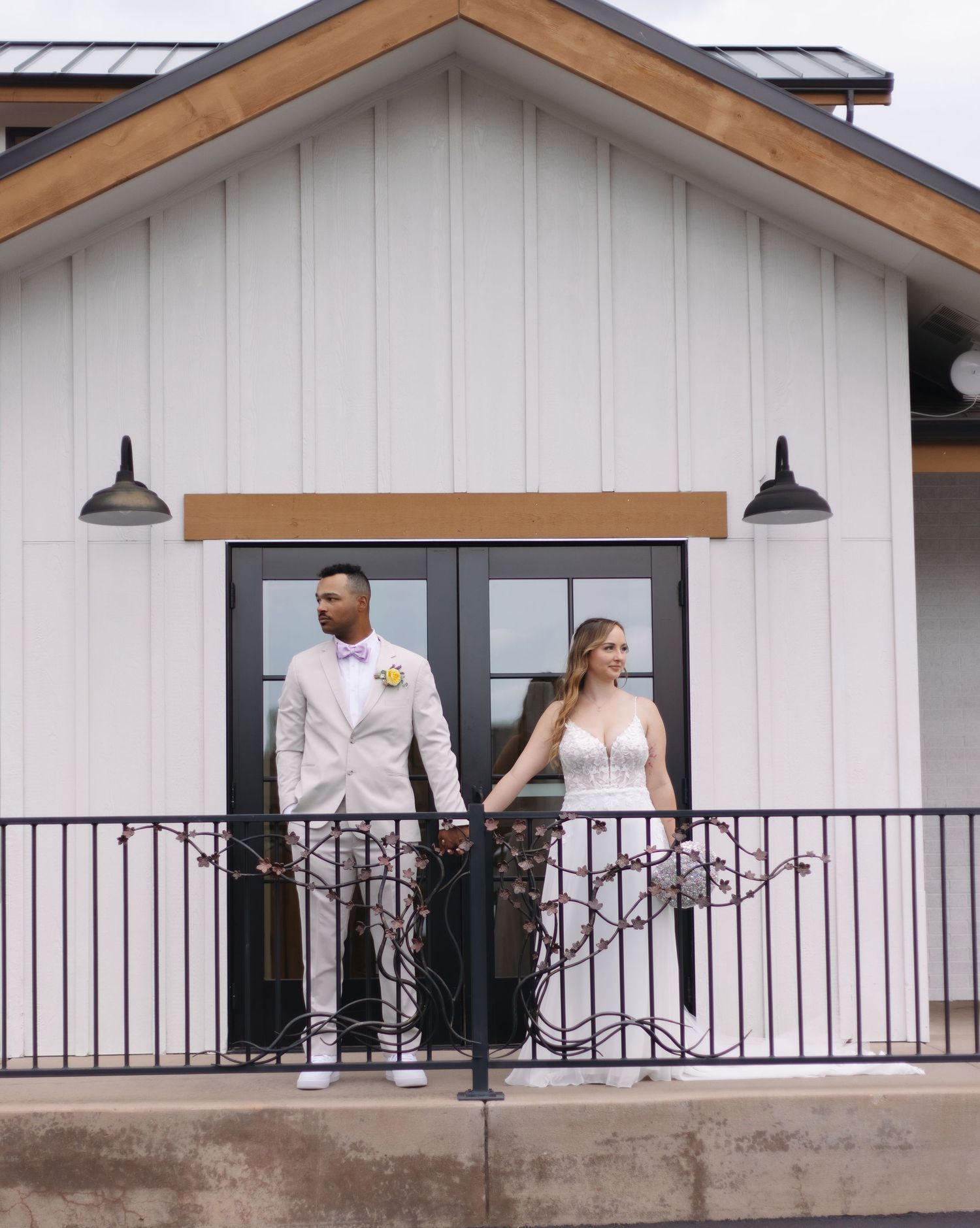 Wedding couple holds hands on a white farmhouse barn balcony, groom in tan suit, bride in lace wedding dress.