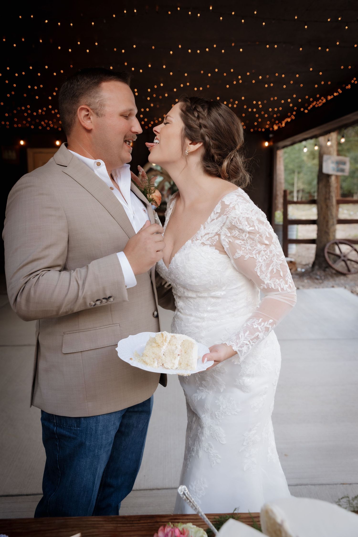 Bride and groom laughing together during wedding cake cutting under warm string lights.