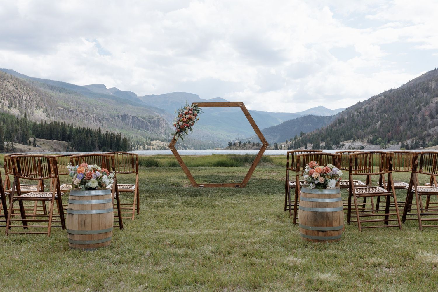 Outdoor wedding ceremony setup with hexagonal arch, wine barrel stands, and mountain lake backdrop.