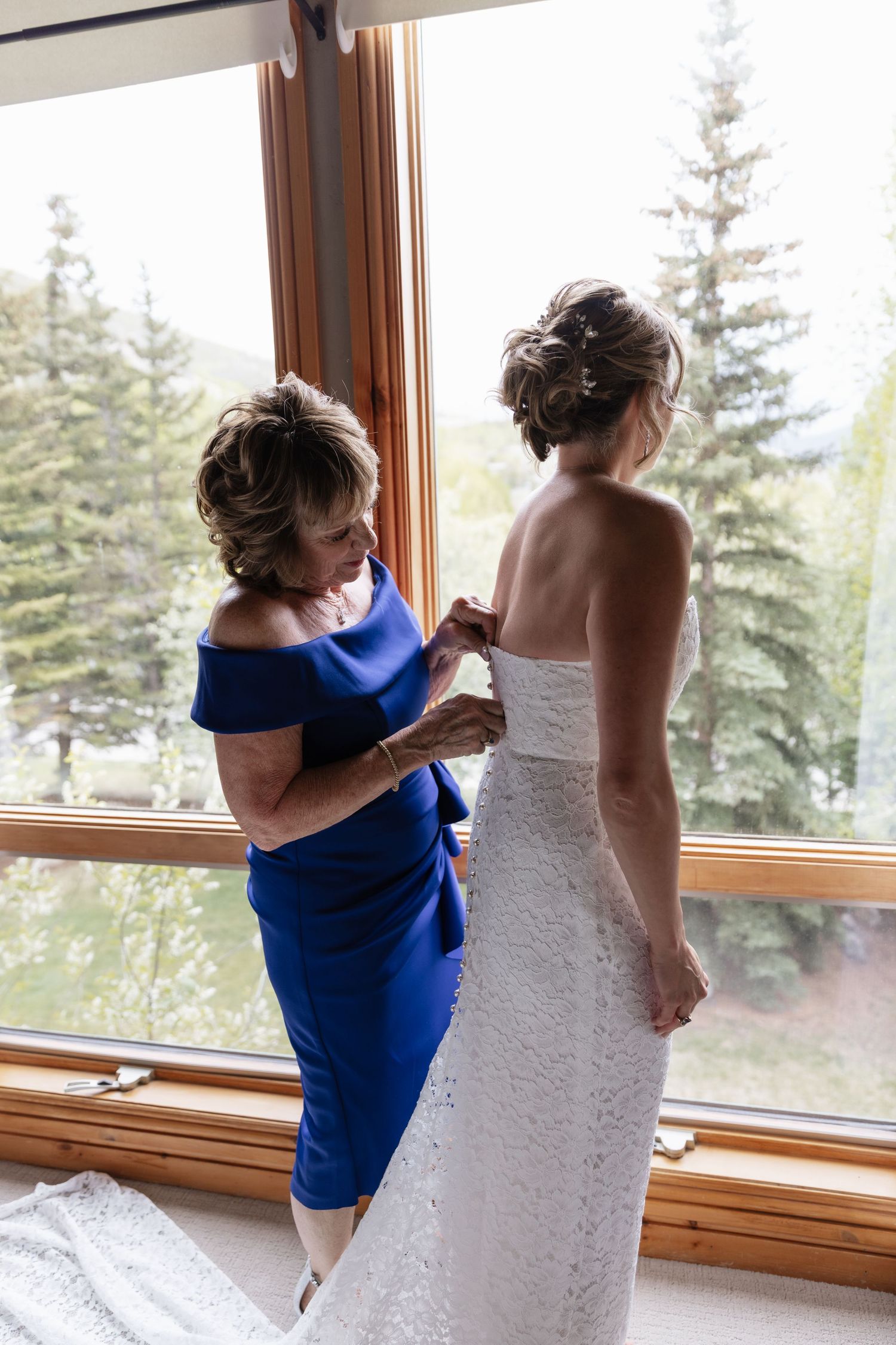 Mother in blue dress buttoning bride's white lace wedding gown by large windows with pine tree views.