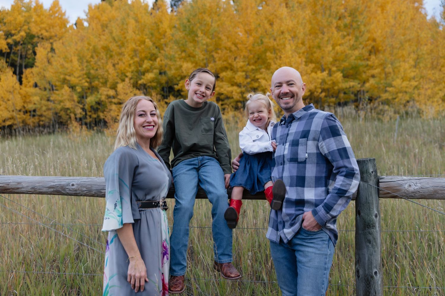 Family of four posing on a wooden fence with golden autumn foliage in the background.