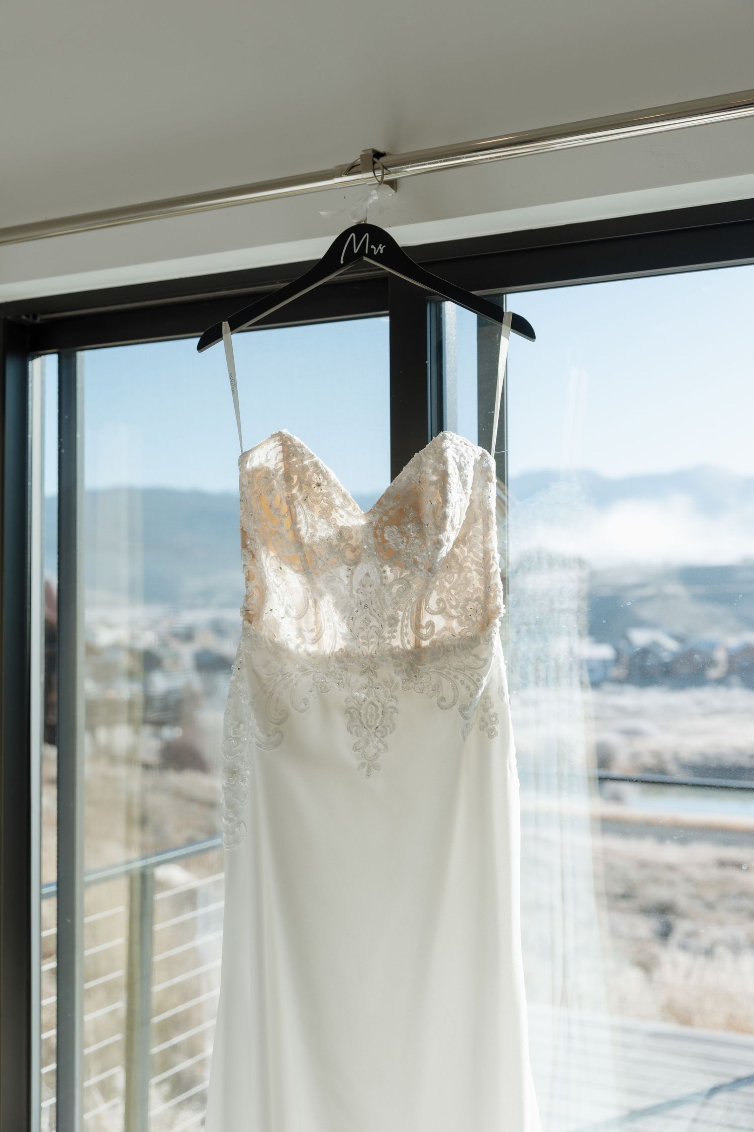 Elegant lace and satin wedding dress hanging on Mrs. hanger against mountain view window.