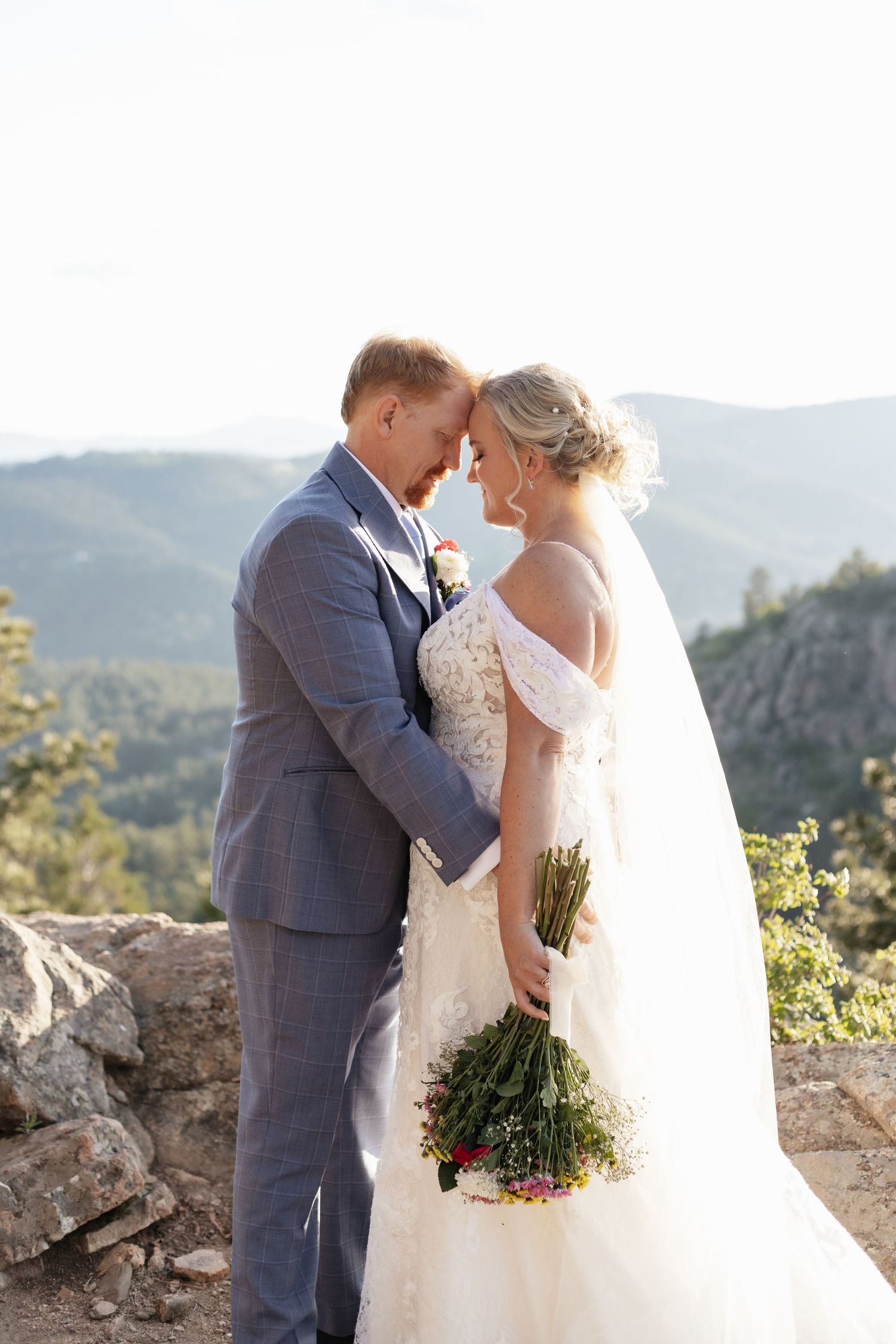 Bride and groom touching foreheads romantically on a rocky mountain overlook with green valley views.