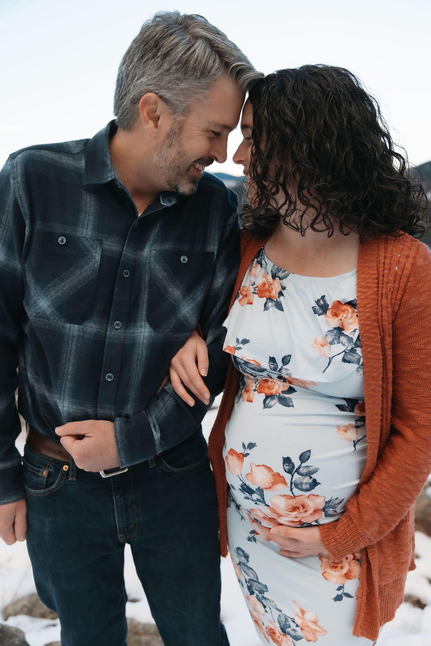 Smiling expectant couple touching foreheads tenderly outdoors in a snowy mountain setting.