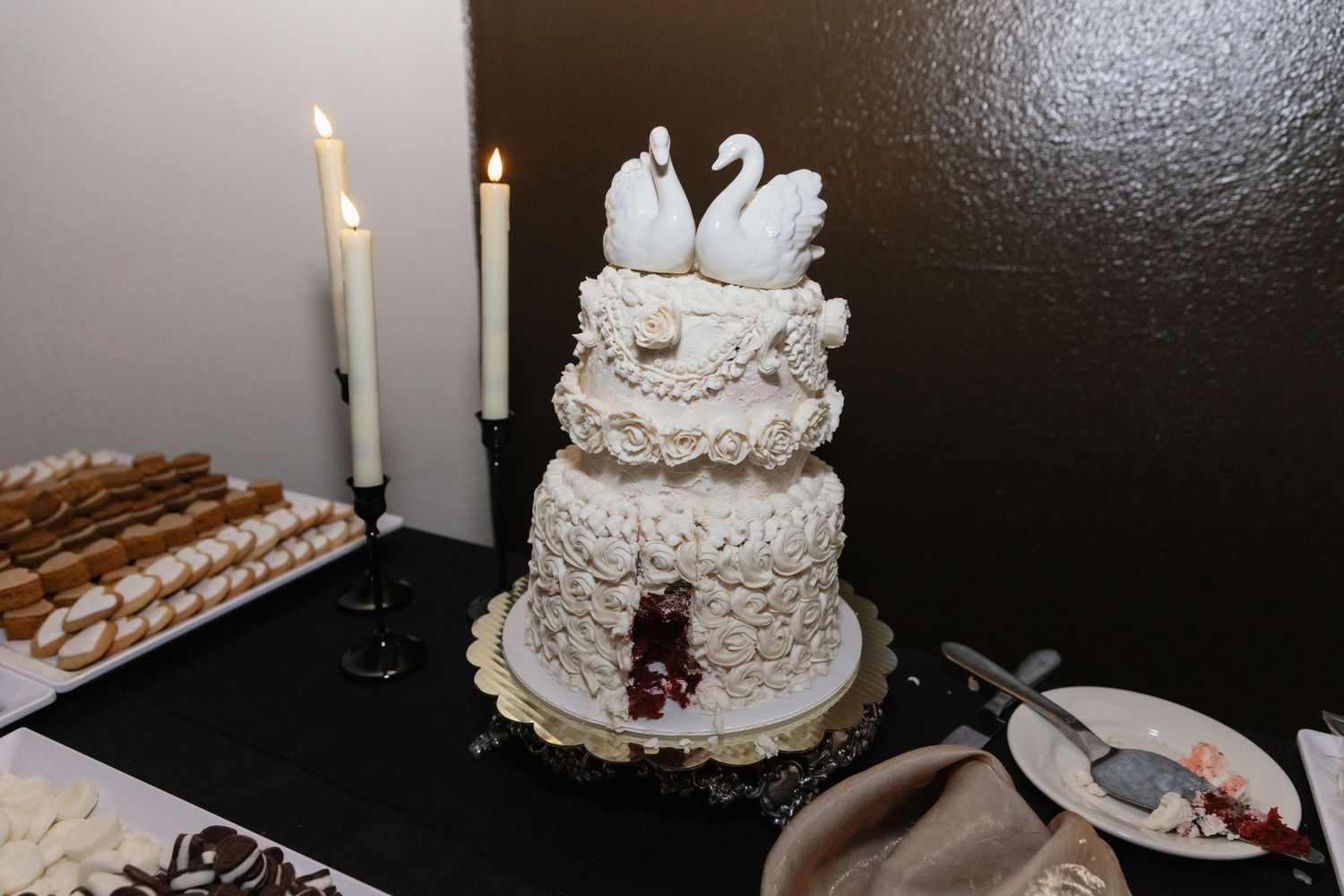 Elegant three-tier white wedding cake topped with swan figurines displayed on decorated dessert table.