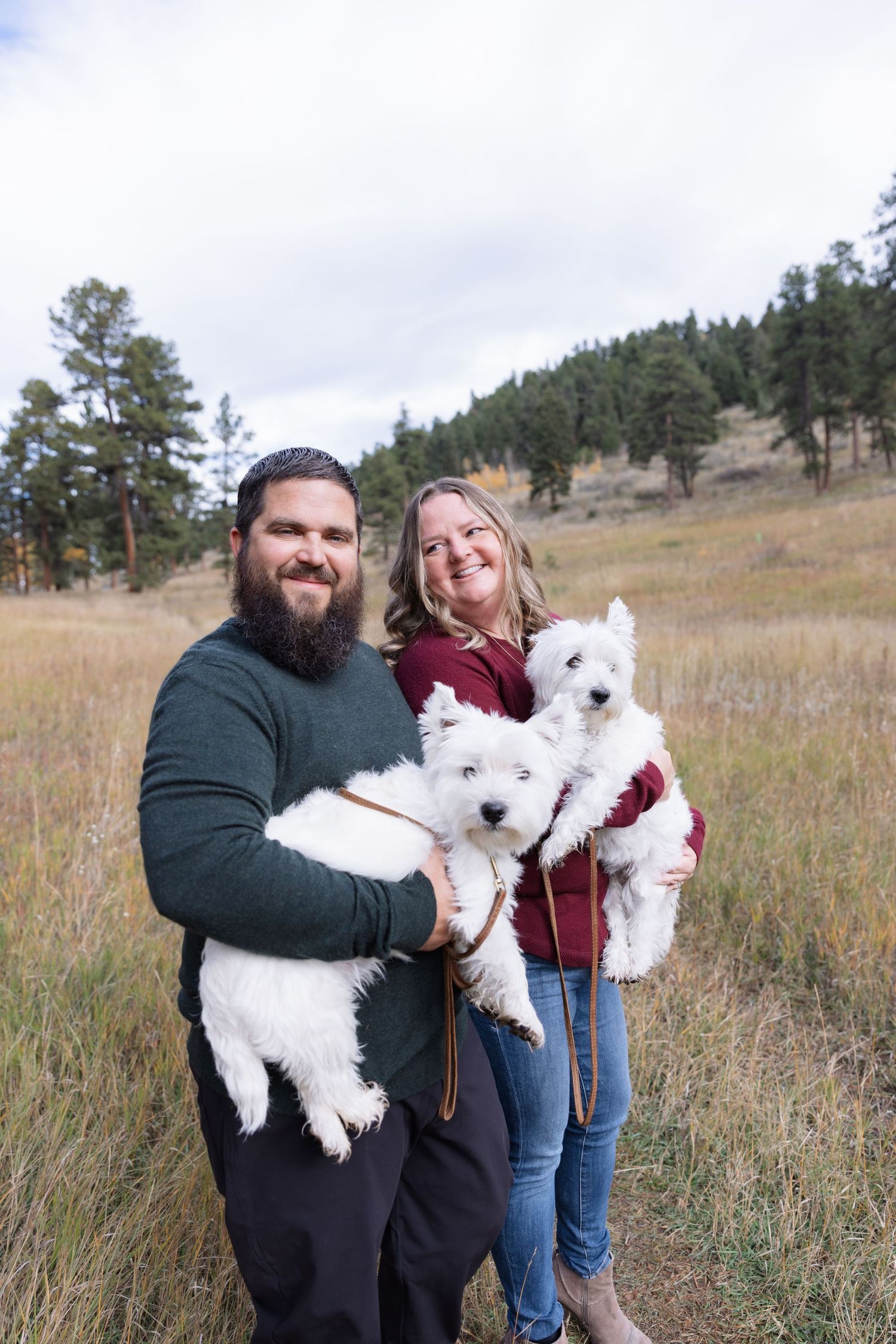 Smiling couple holding two fluffy white puppies in an autumn mountain meadow.