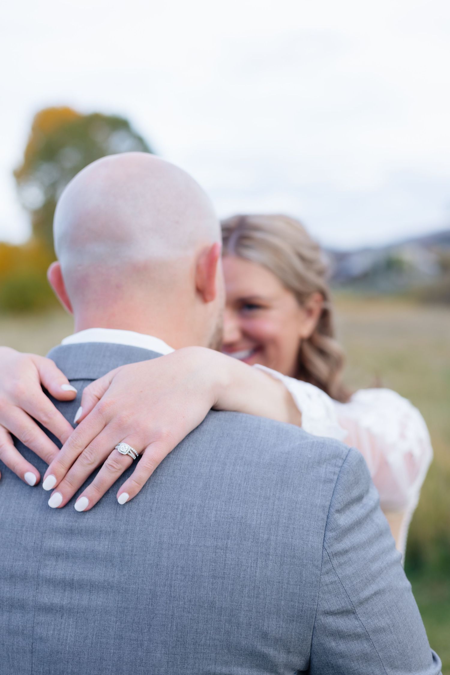 Smiling bride embraces bald groom from behind in autumn field, showcasing her diamond wedding ring on his shoulder.