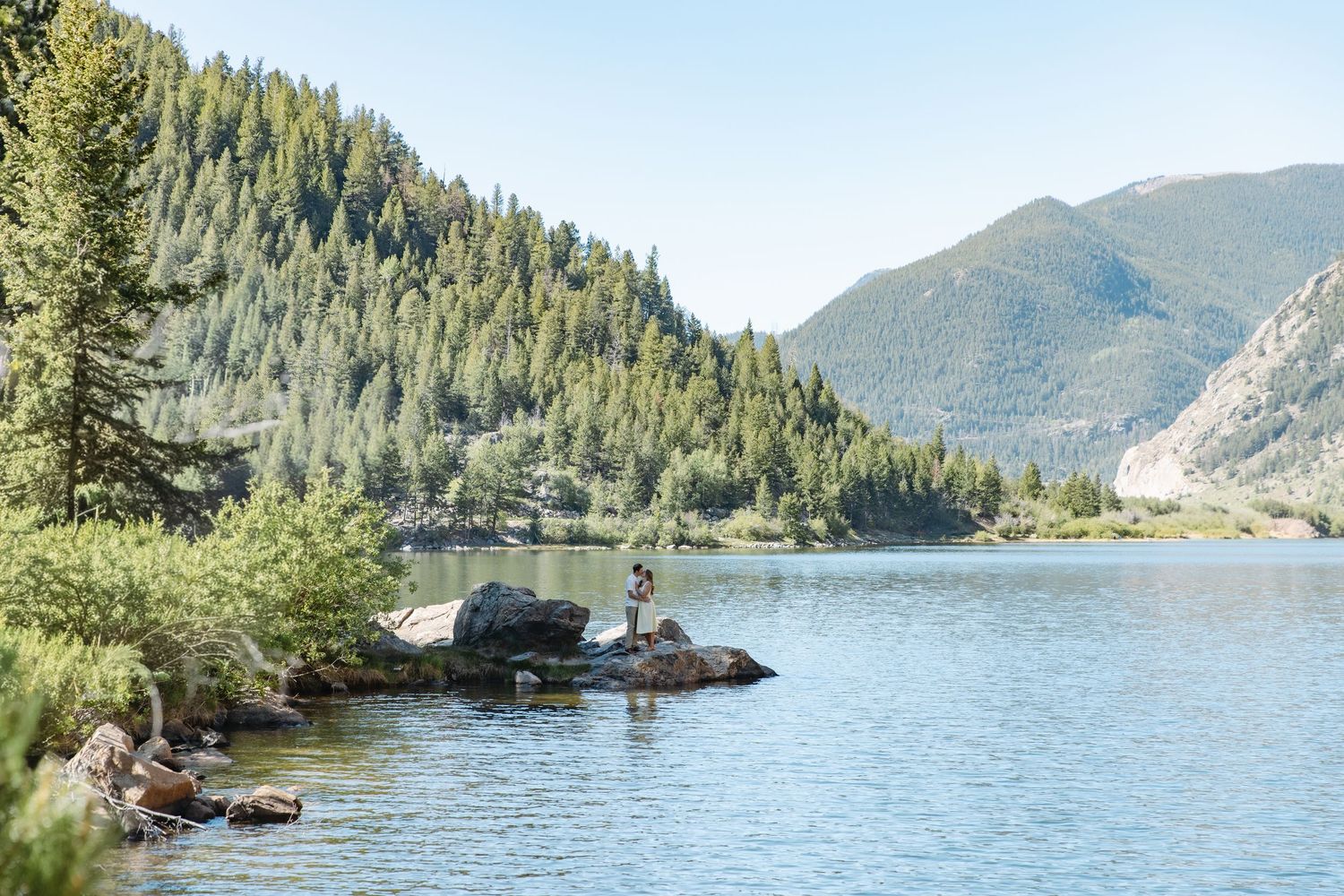 Person standing on rocks fishing in a serene mountain lake surrounded by pine trees and scenic hills on a clear day.