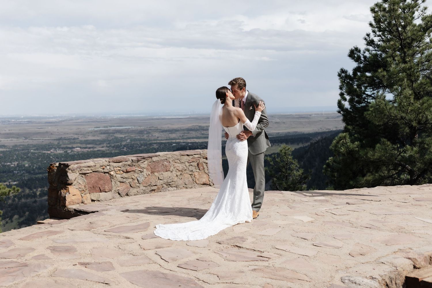 Bride and groom share a kiss on a rocky overlook with sweeping valley views and pine trees at a Colorado wedding.