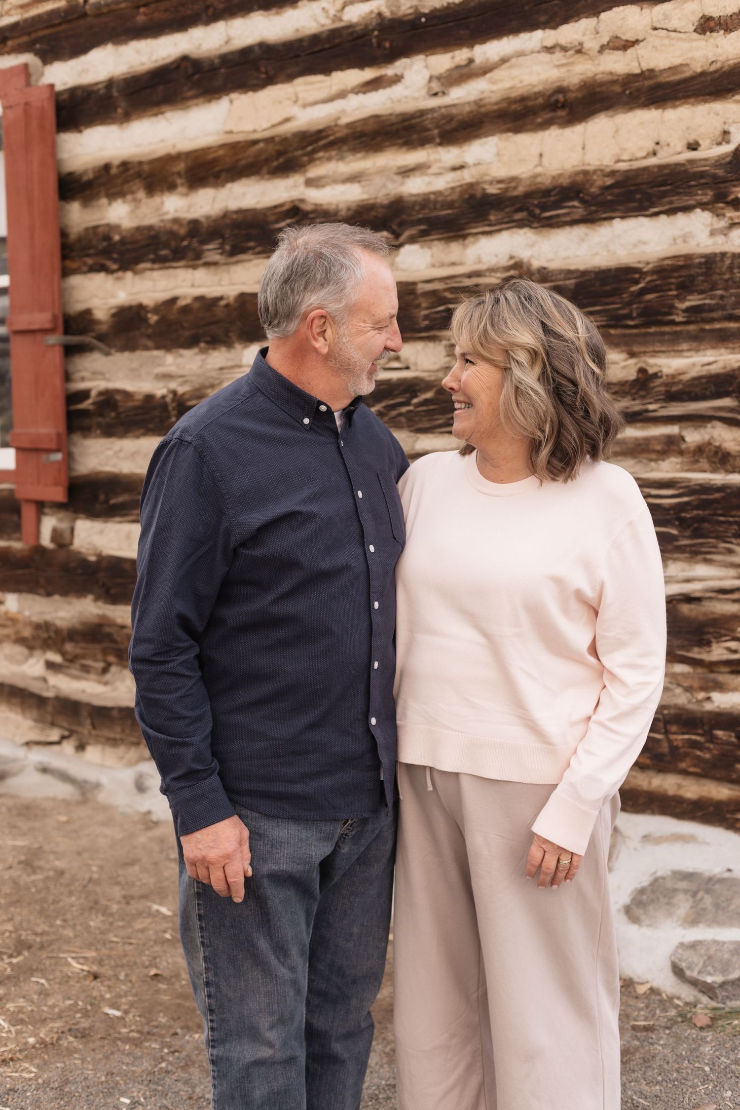 Smiling mature couple sharing a tender moment outside a rustic log cabin wall.