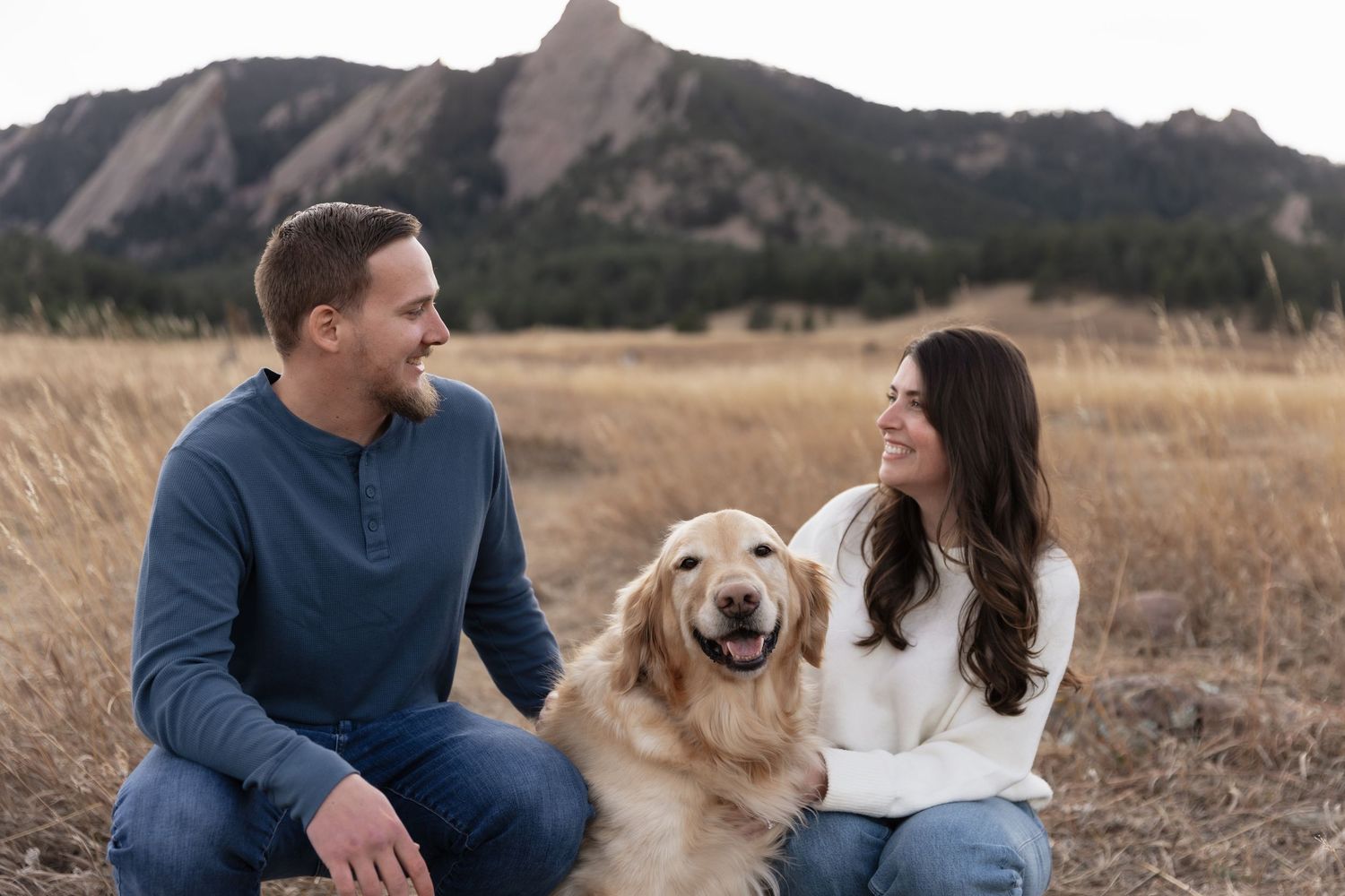 Smiling couple sits with golden retriever in Colorado Flatirons field.