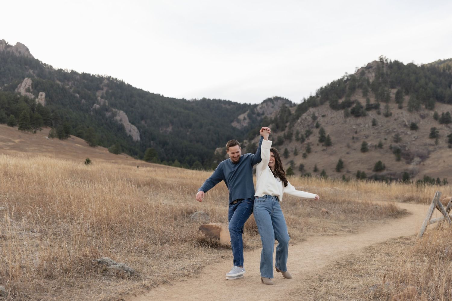 Couple laughs and dances on mountain trail in dry grass field.