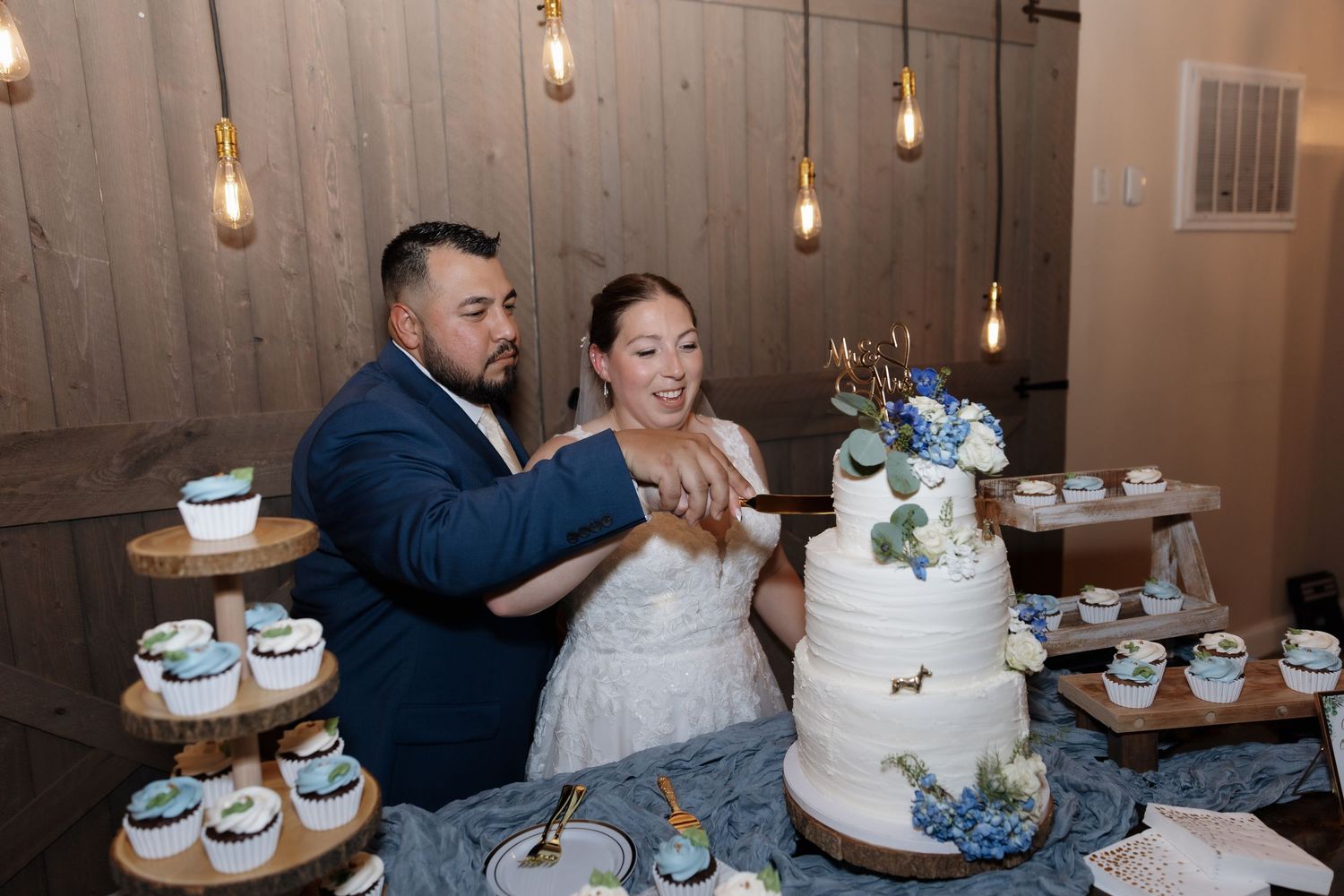 Bride and groom cutting white wedding cake with cupcakes at rustic barn reception with Edison bulb lighting.