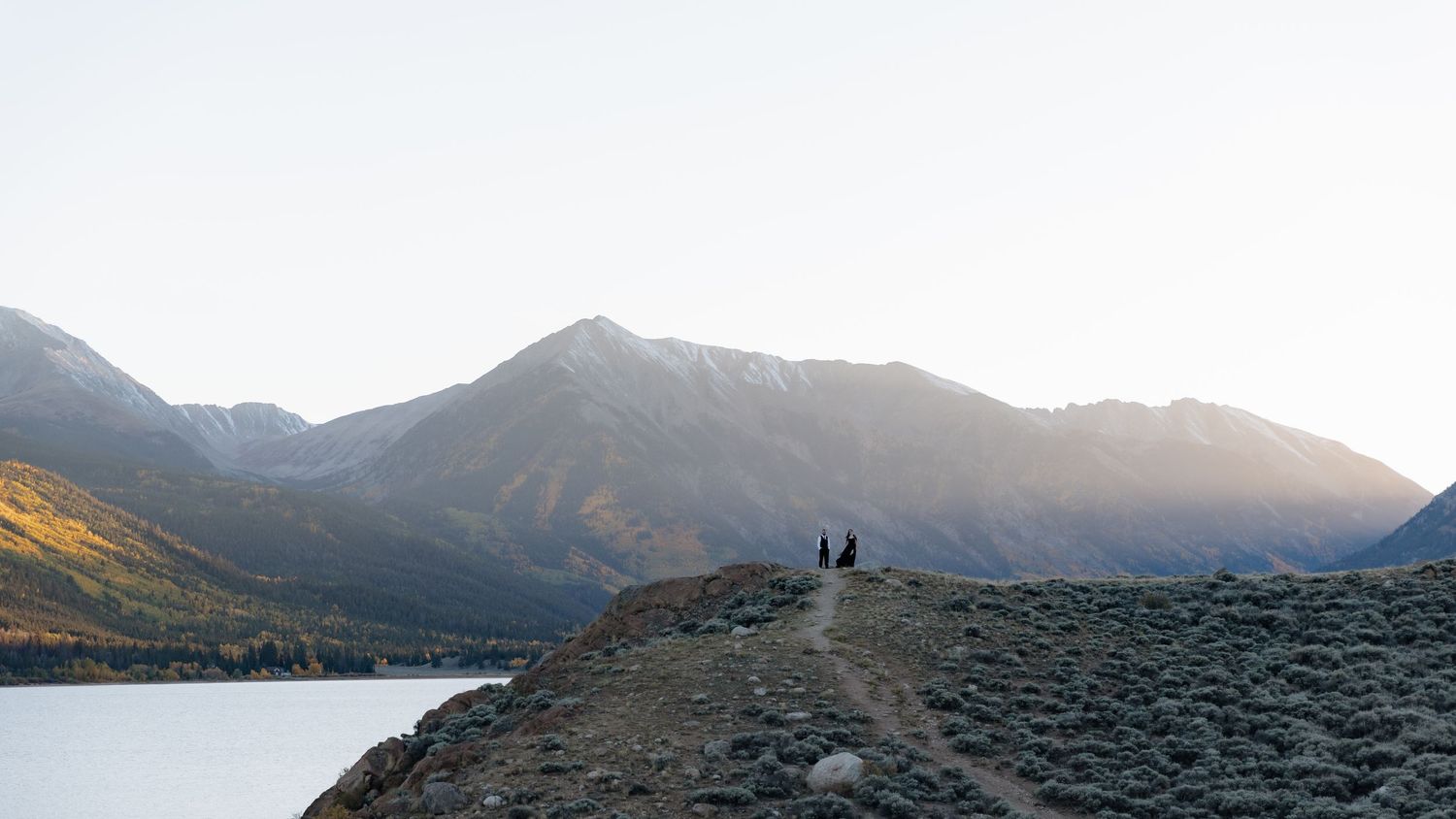 Two figures standing on rocky overlook above a serene lake with majestic mountain backdrop at sunset. Twin Lakes