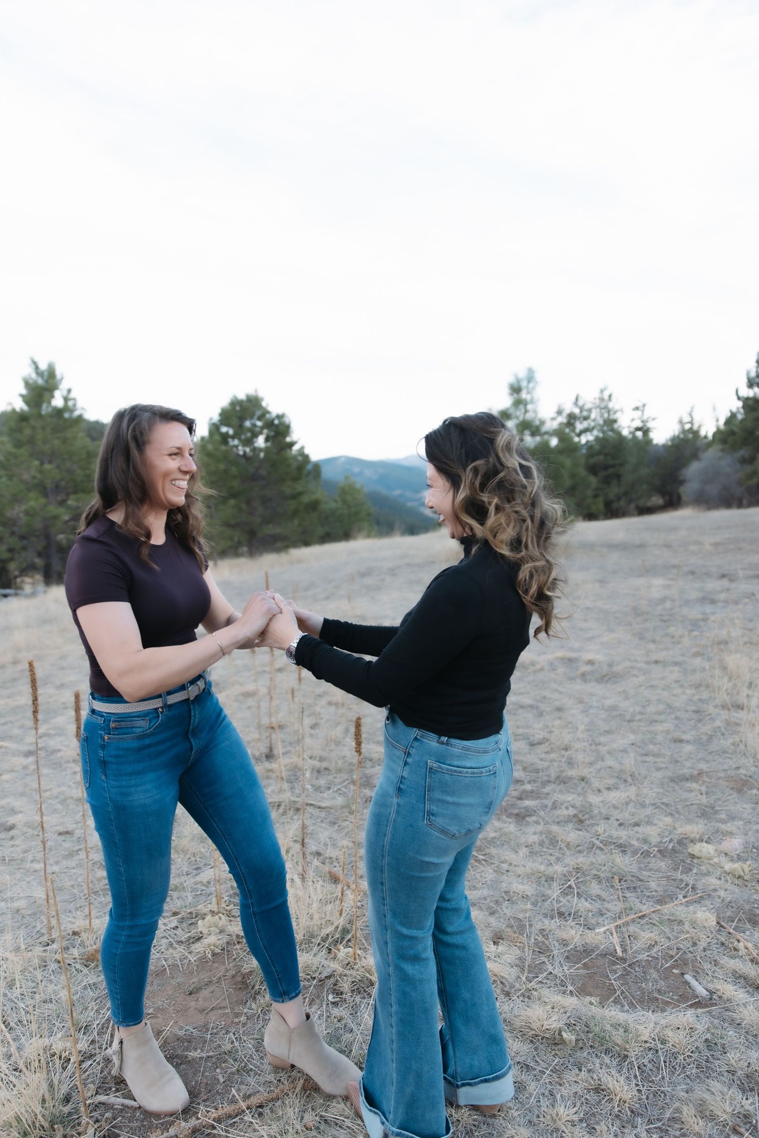 Two women in black tops and jeans holding hands and laughing together in an open field.