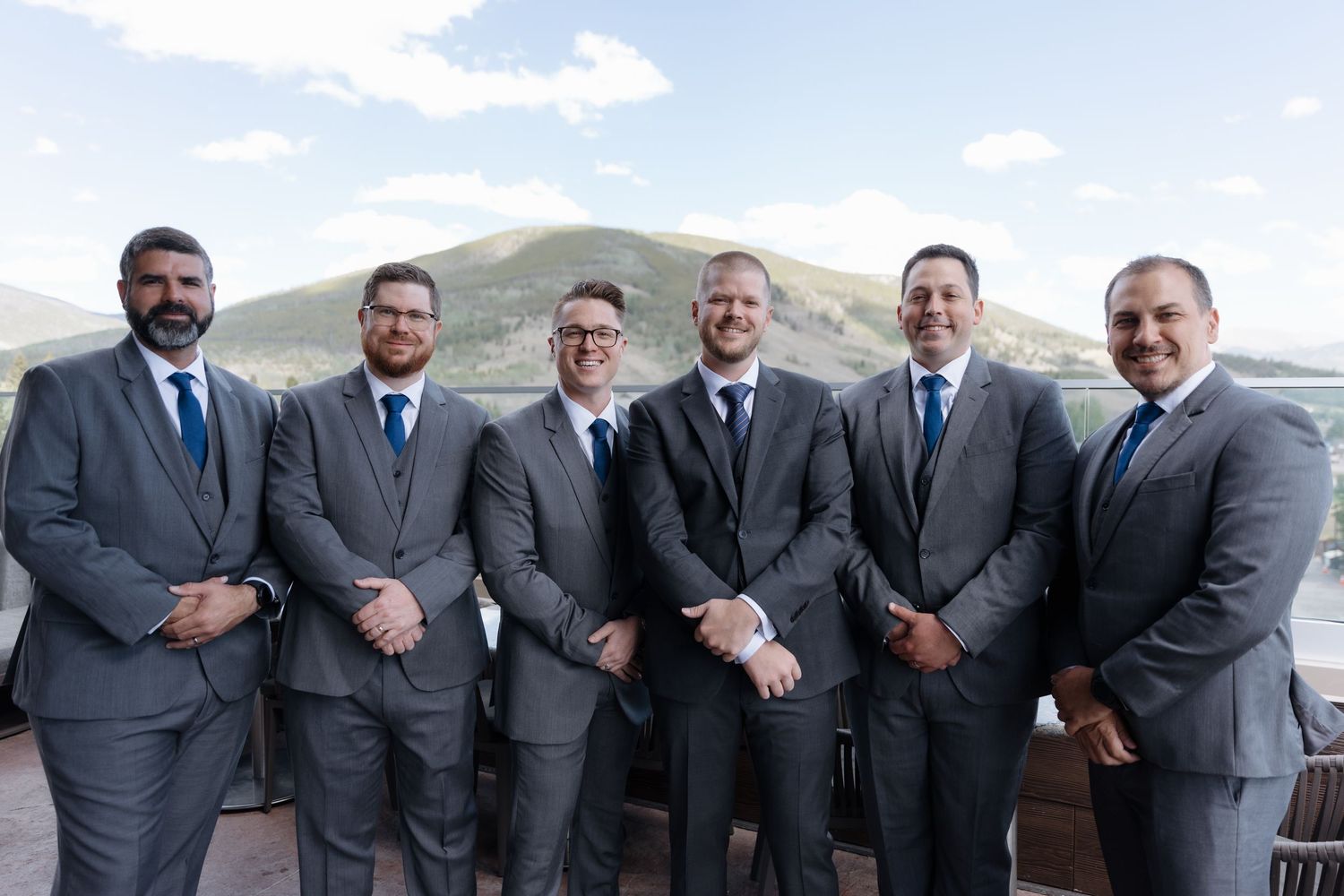 Six groomsmen in matching gray suits with blue ties pose outdoors with a scenic mountain backdrop on a wedding day.