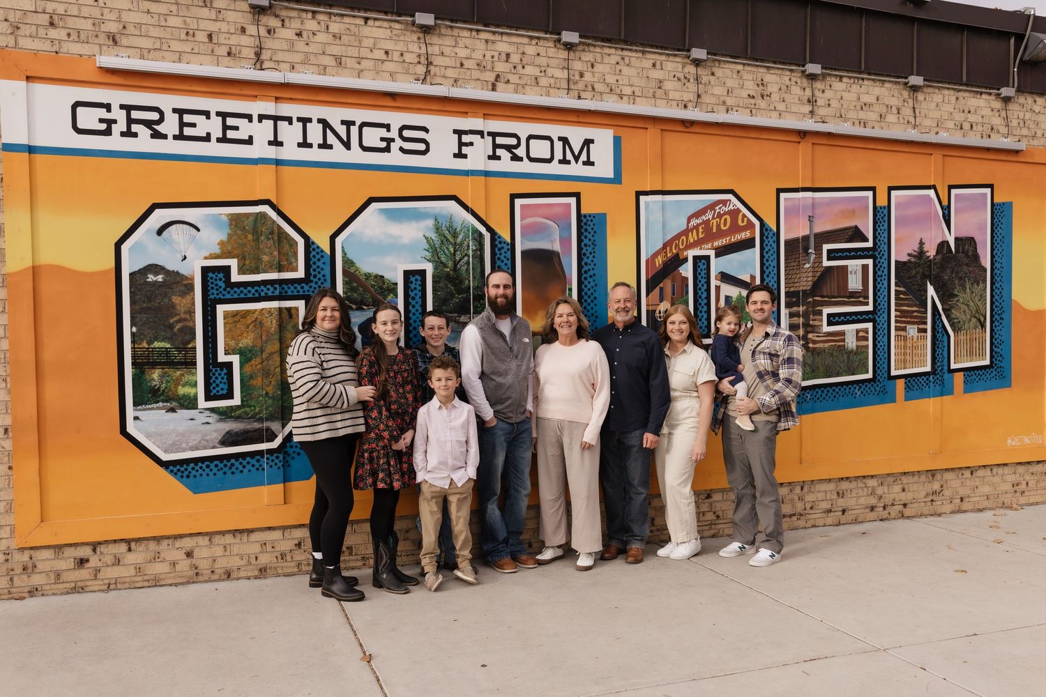 Large extended family posing in front of a colorful Greetings from Golden mural outdoors.