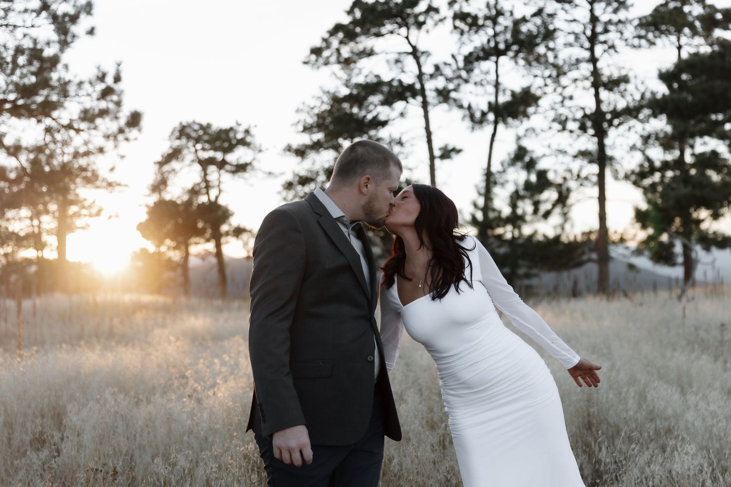 Bride and groom sharing a sunset kiss in a golden meadow surrounded by pine trees.