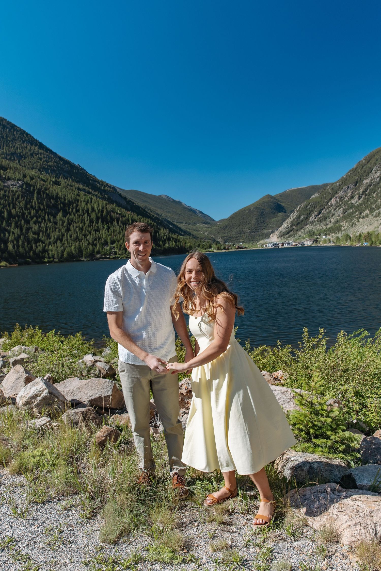 Couple holding hands smiling by a mountain lake with green hills and clear blue sky in background.