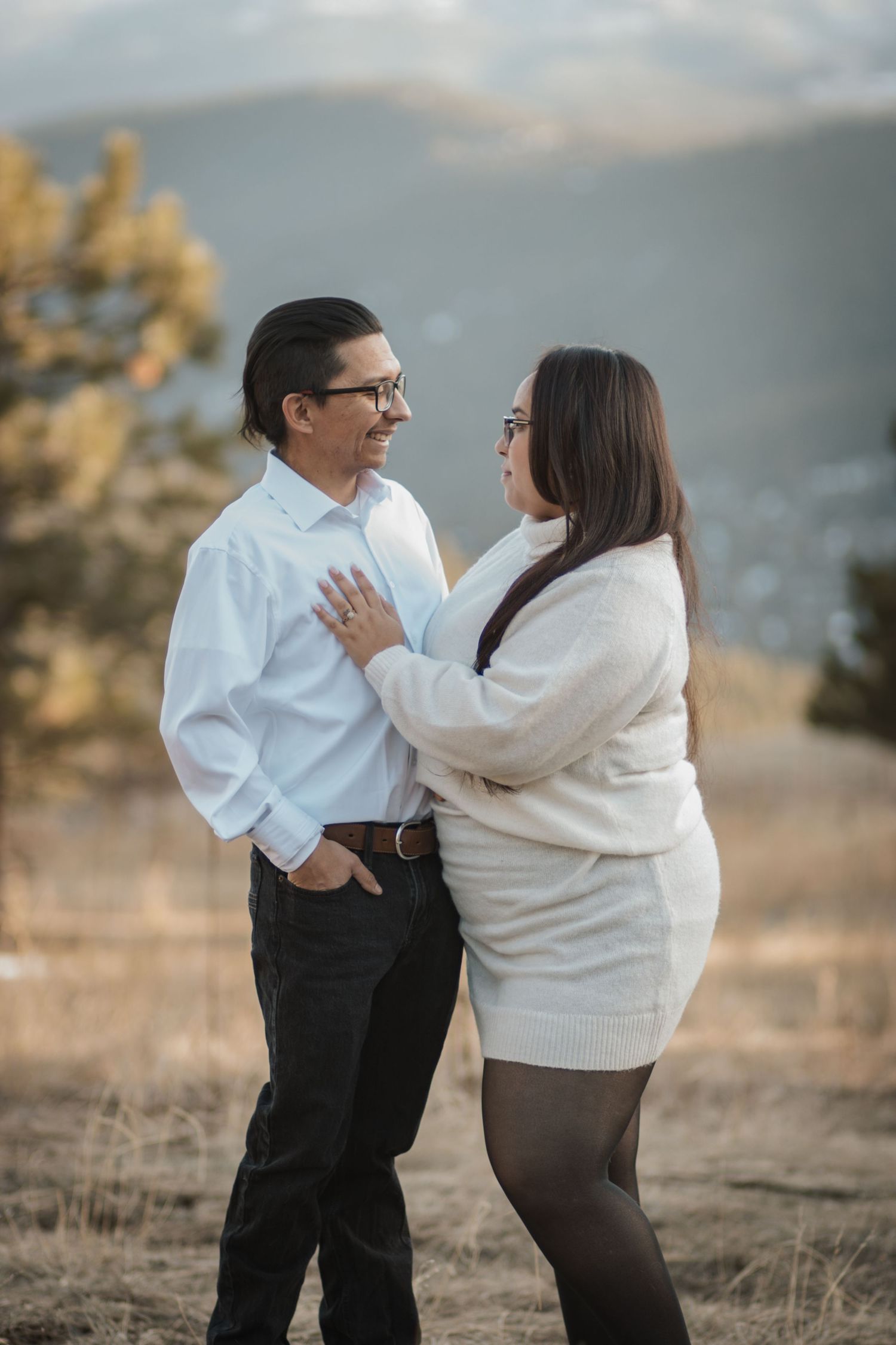 Smiling couple embracing in a golden field with mountain backdrop during engagement shoot.