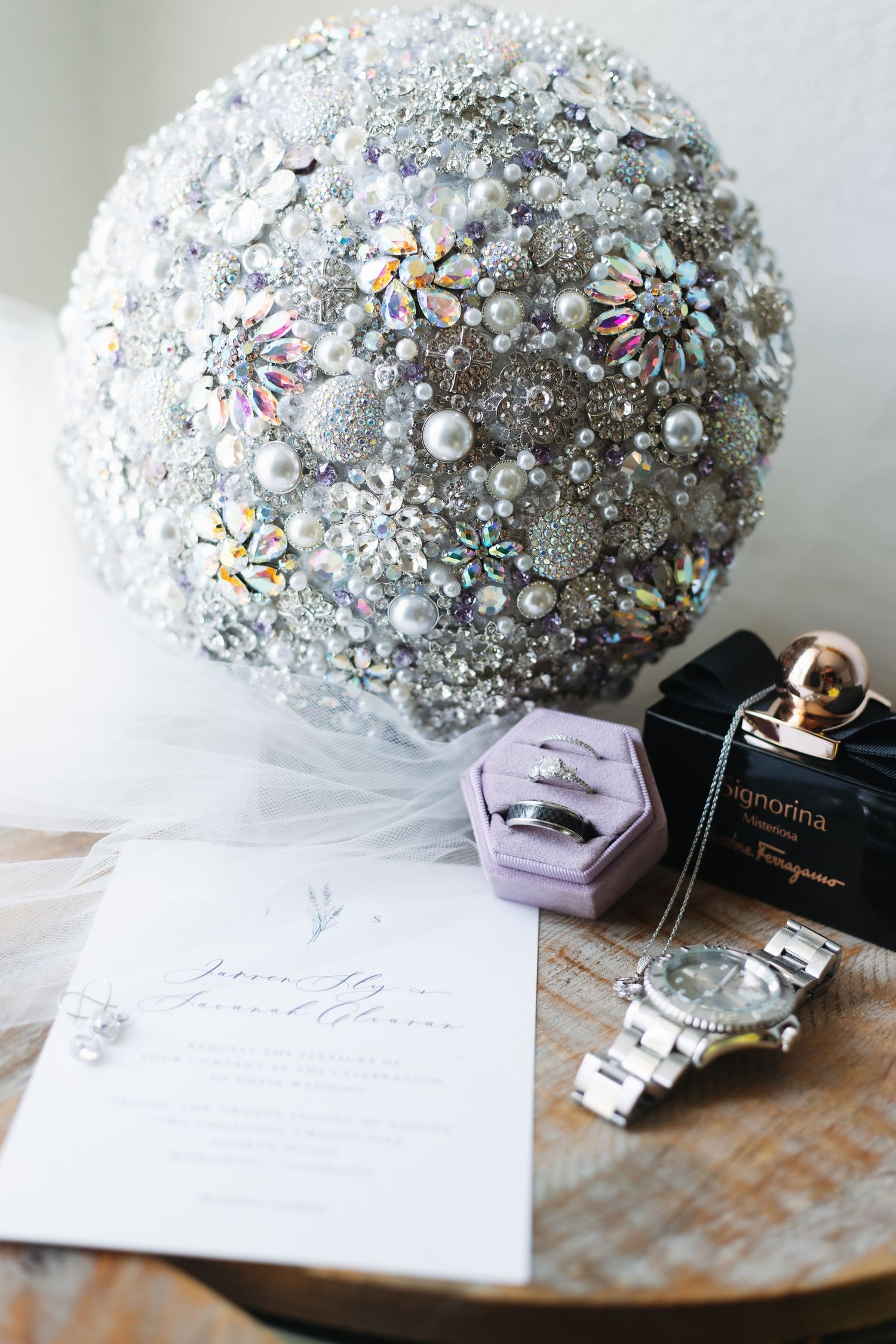 Elegant brooch bouquet of pearls and crystals displayed with wedding rings, watch, perfume, and invitation on wood table.