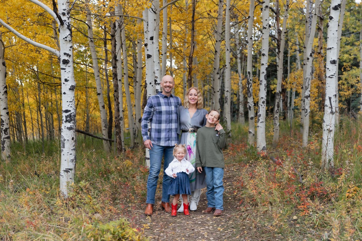 Family of four posing among white aspen trees with golden autumn foliage on a forest trail.