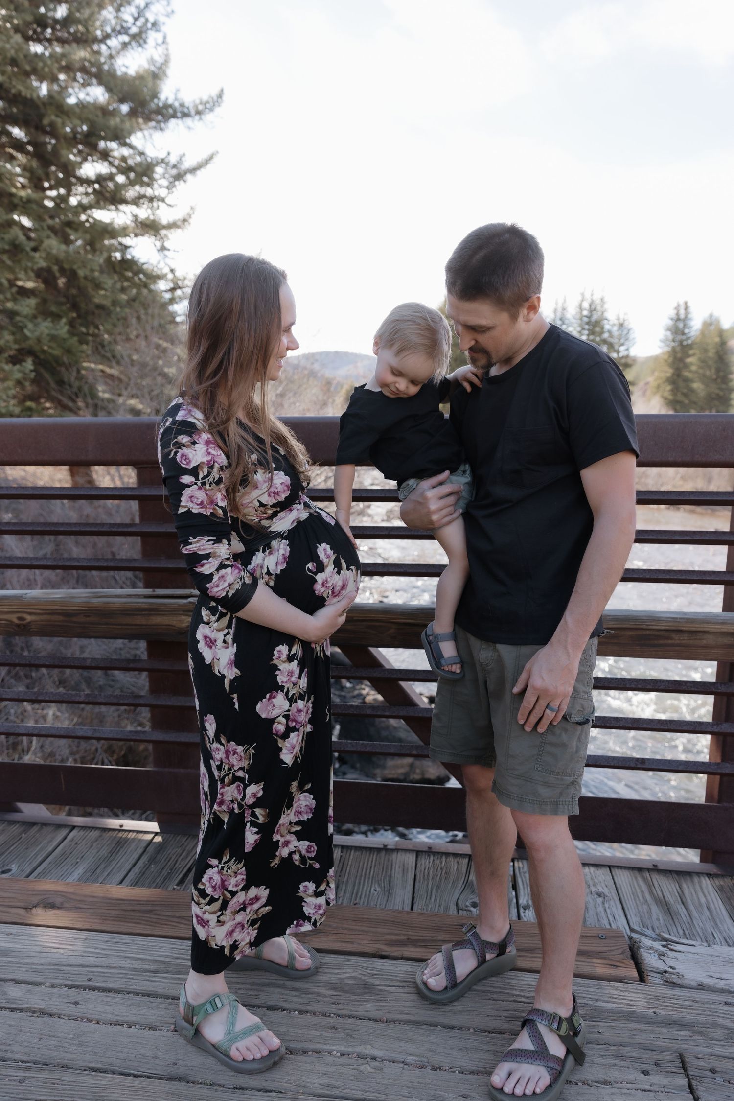 Pregnant woman and man holding toddler on a wooden bridge surrounded by pine trees.