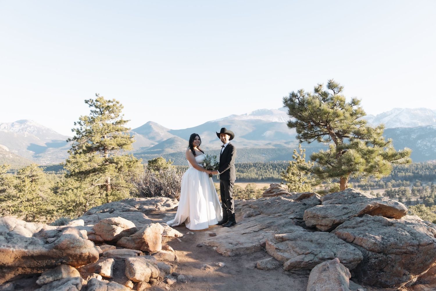 Couple exchanges vows on a rocky mountain summit at sunrise, bride in white gown, groom wearing a black cowboy hat.
