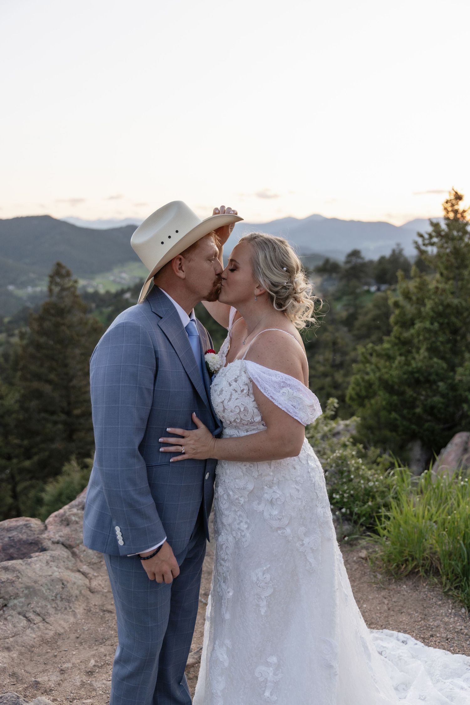 Couple shares a kiss at sunset with mountain views, groom wearing a cowboy hat and blue suit at their wedding.