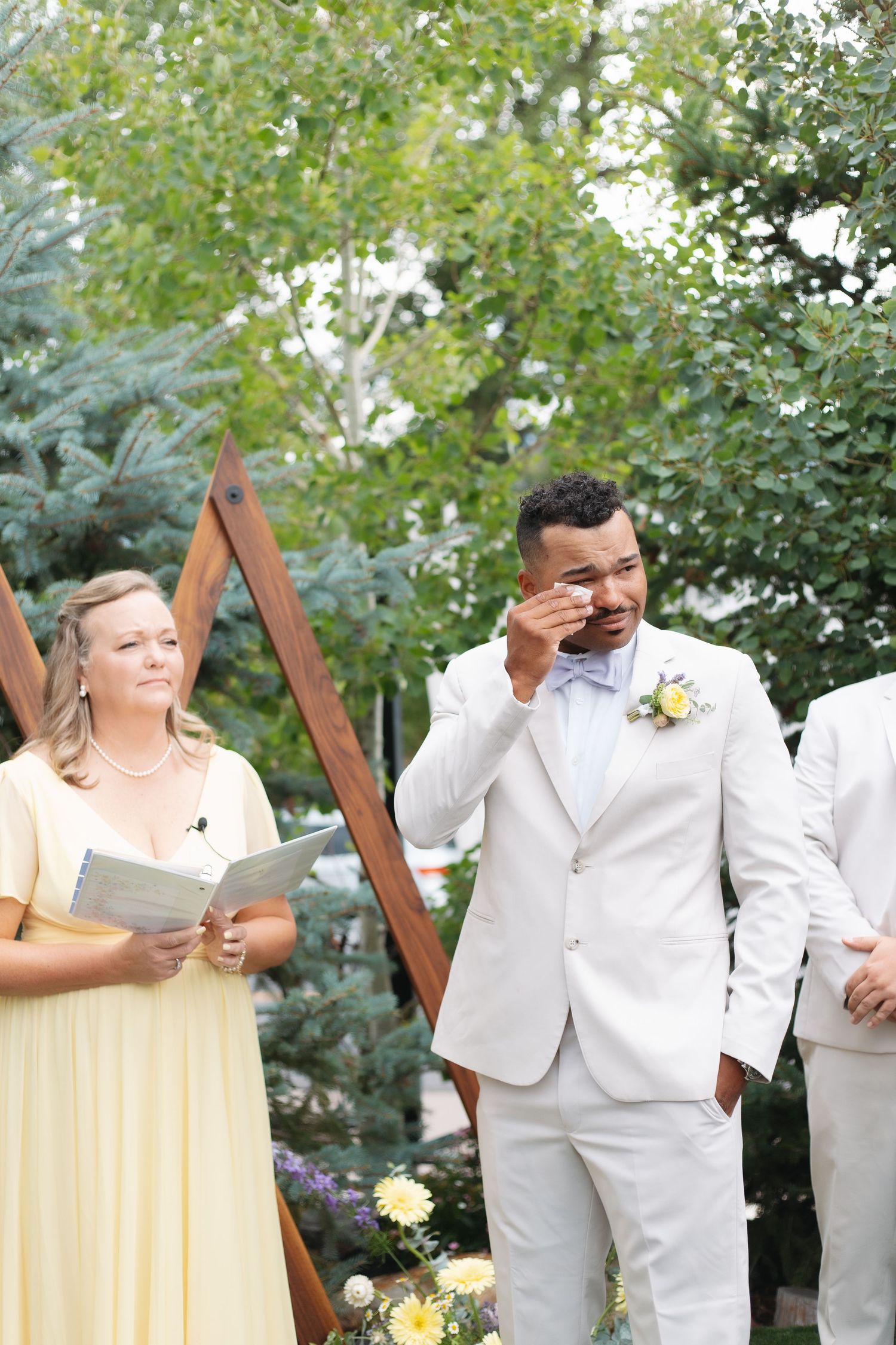 Emotional groom in white suit wipes tears during outdoor wedding ceremony.