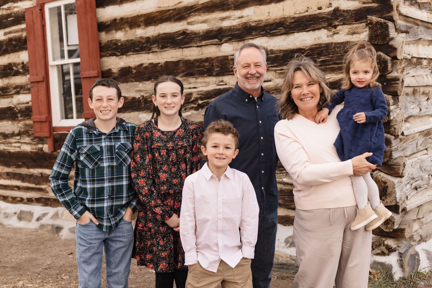 Happy extended family of six posing and smiling in front of a historic log cabin.
