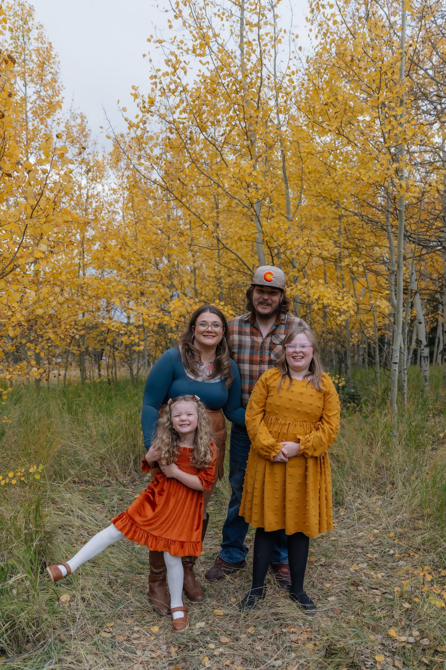 Family of four with two girls in autumn dresses posing in front of vibrant yellow aspen trees.