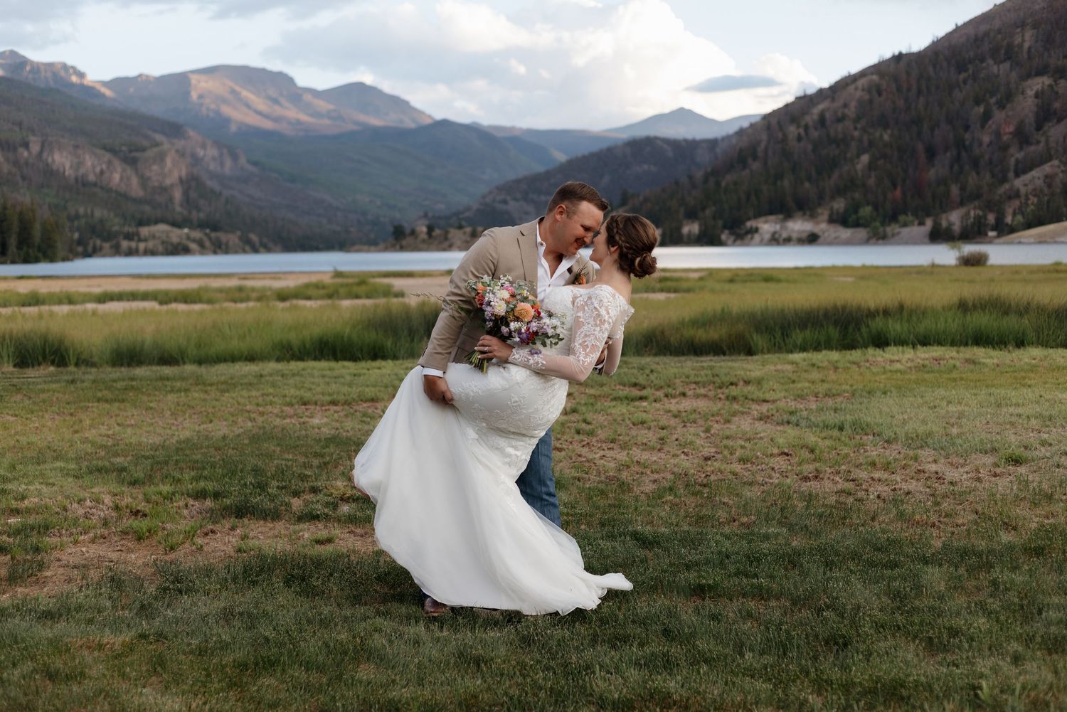 Bride and groom share romantic dip kiss in mountain meadow with lake and peaks backdrop on wedding day.