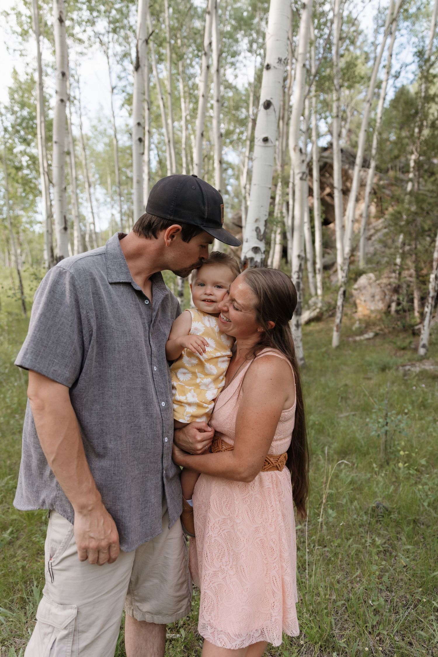 Young family with baby sharing kisses among white aspen trees in a lush green forest.