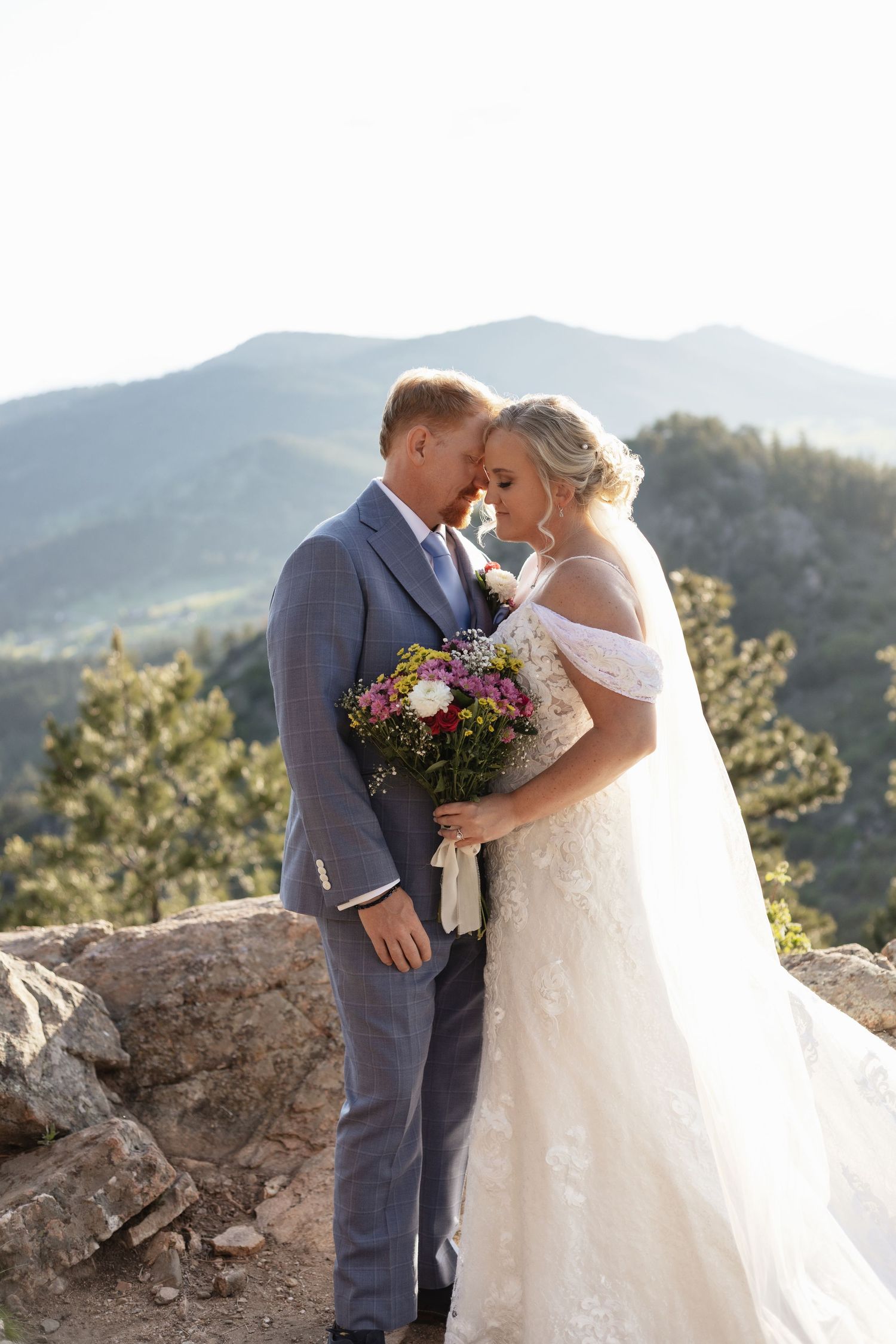 Bride and groom share a romantic forehead touch on a mountain overlook, bride holding a colorful bouquet in wedding attire.
